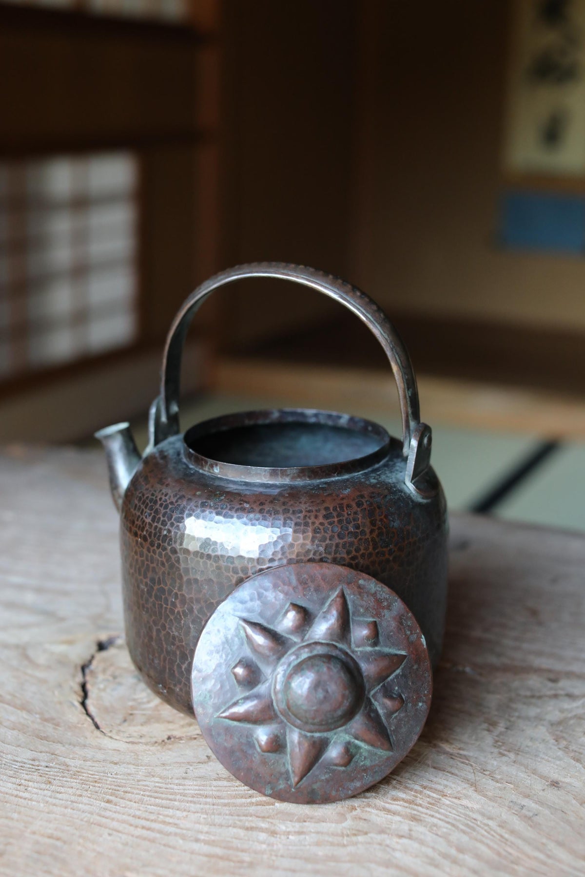 An antique copper teapot with a decorative lid sits on a wooden surface, with a blurred background suggesting an indoor setting.