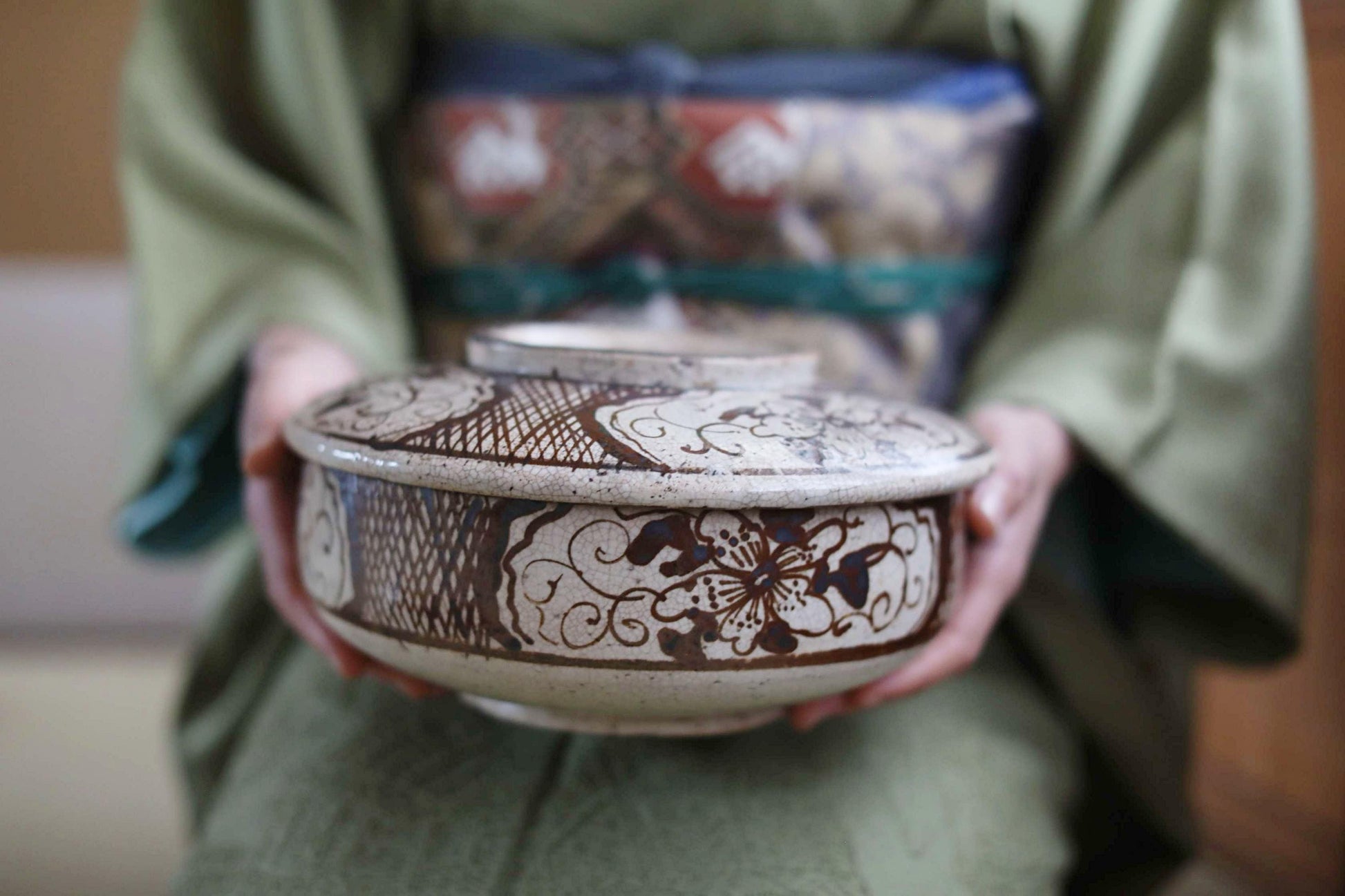 A person wearing a green kimono holds a decorative ceramic bowl with intricate floral patterns.