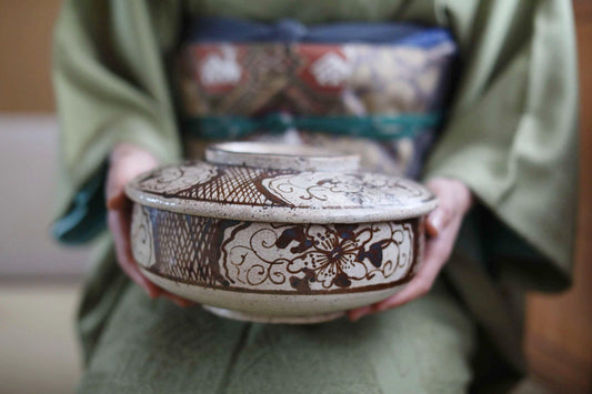 A person wearing a green kimono holds a decorative ceramic bowl with intricate floral patterns.