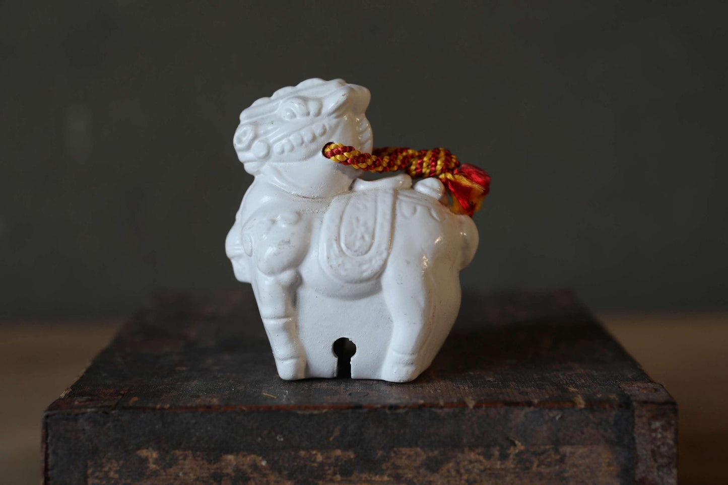 A white ceramic figurine of a lion with a red and yellow rope around its neck is displayed on a wooden surface.