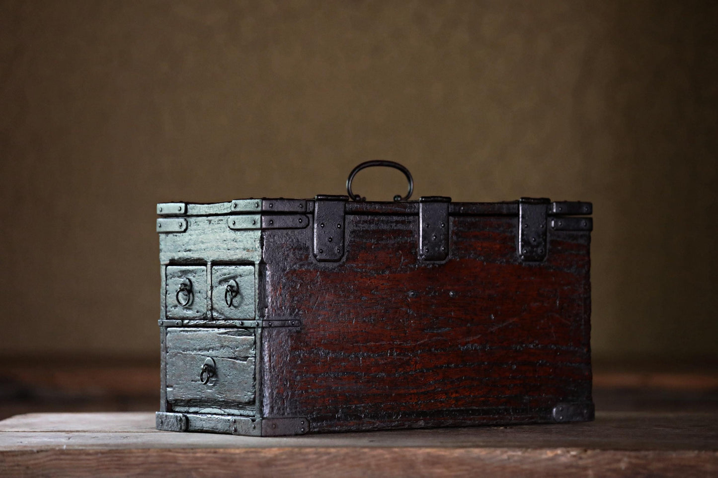 An old, wooden chest with metal clasps sits on a wooden surface against a plain background.