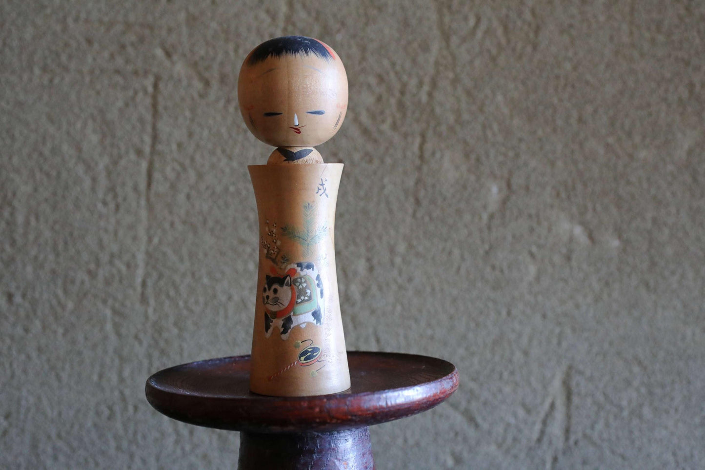 A wooden doll with a face and a cat design sits on a wooden stand against a textured wall.