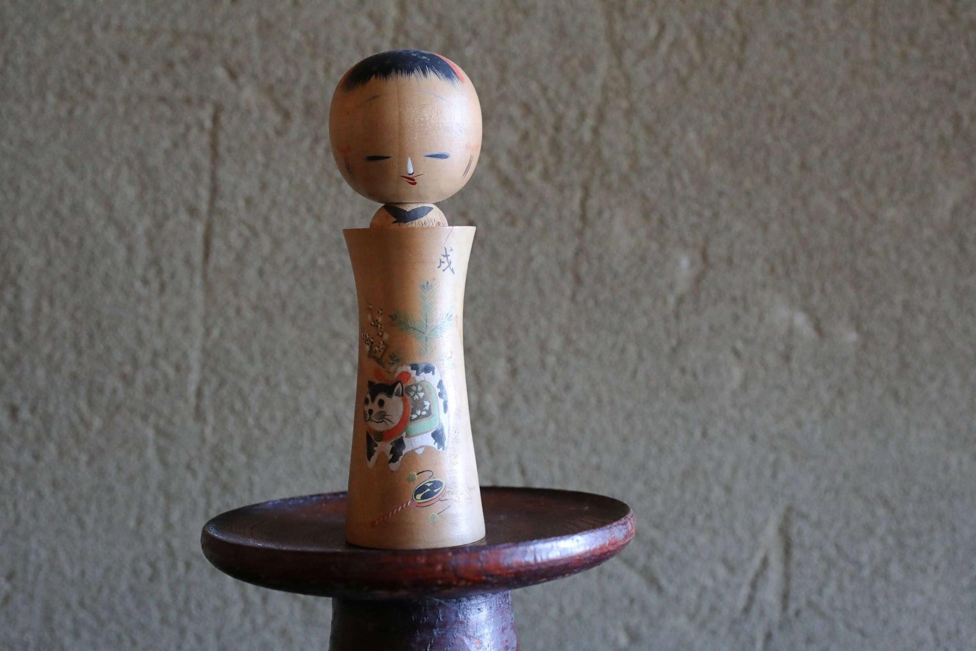 A wooden doll with a face and a cat design sits on a wooden stand against a textured wall.