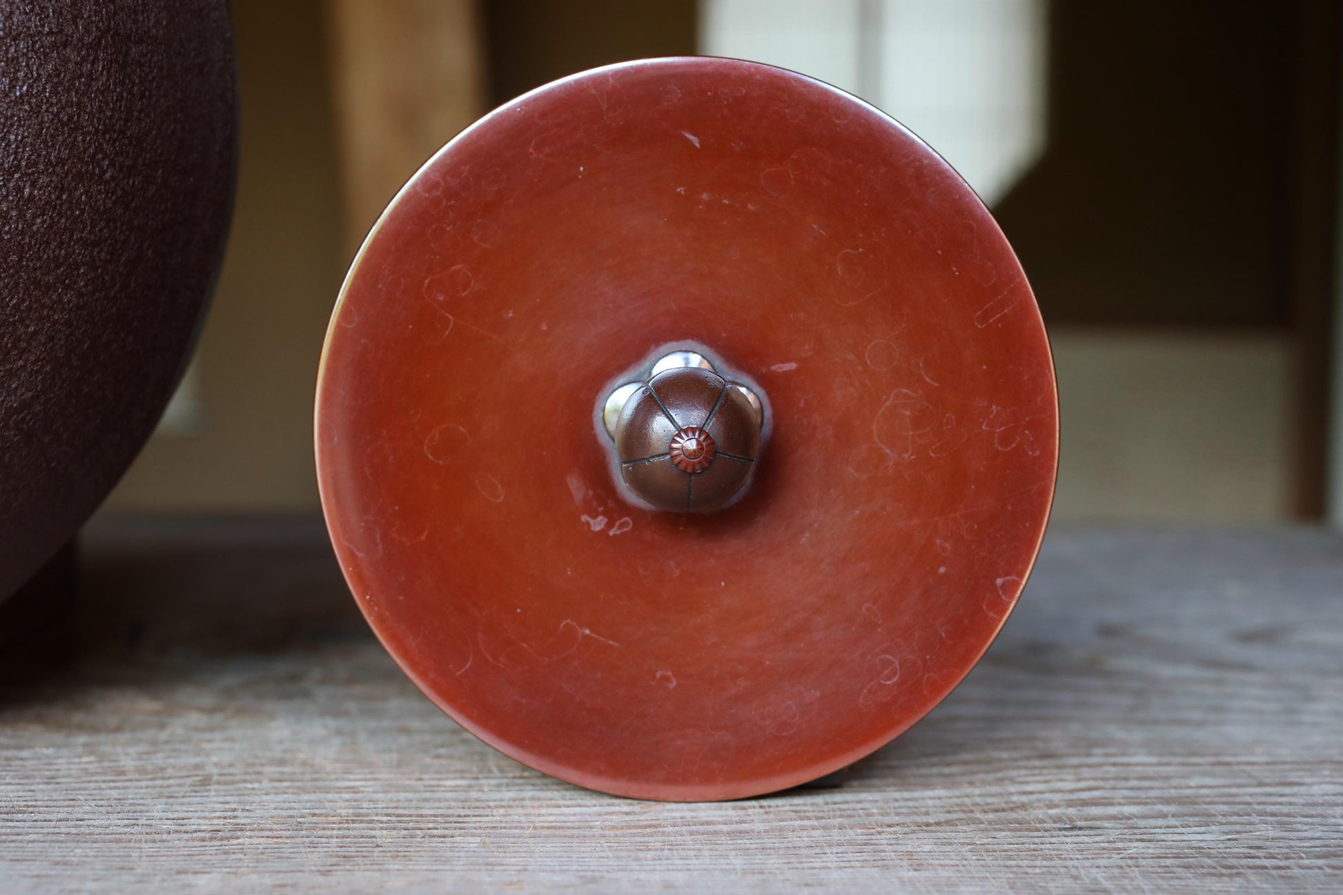 a close-up view of a red ceramic object with a metal flower-shaped knob on it, placed on a wooden surface.