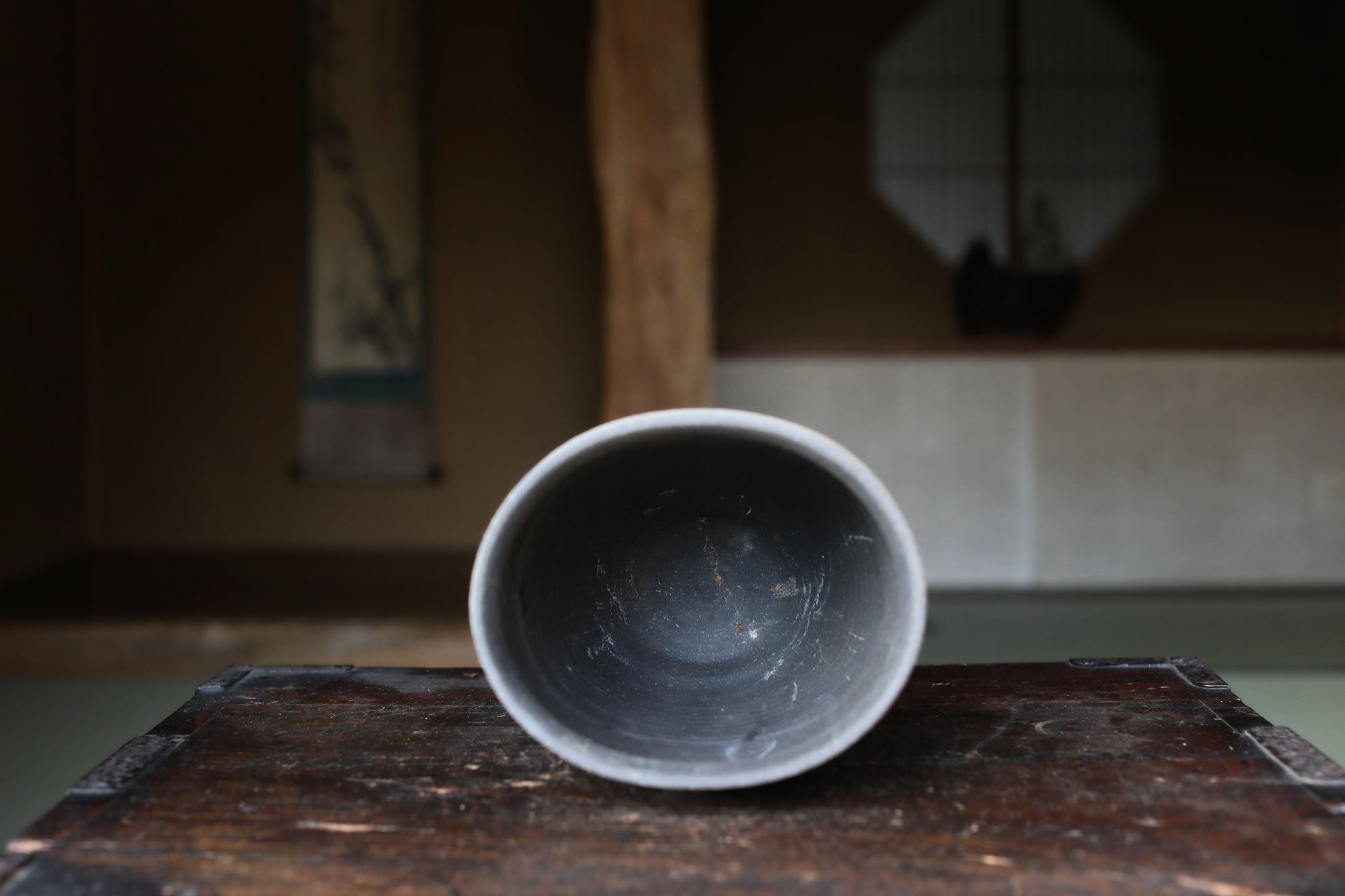 a gray ceramic bowl placed on a wooden surface, with a blurred background that includes a window and a painting.
