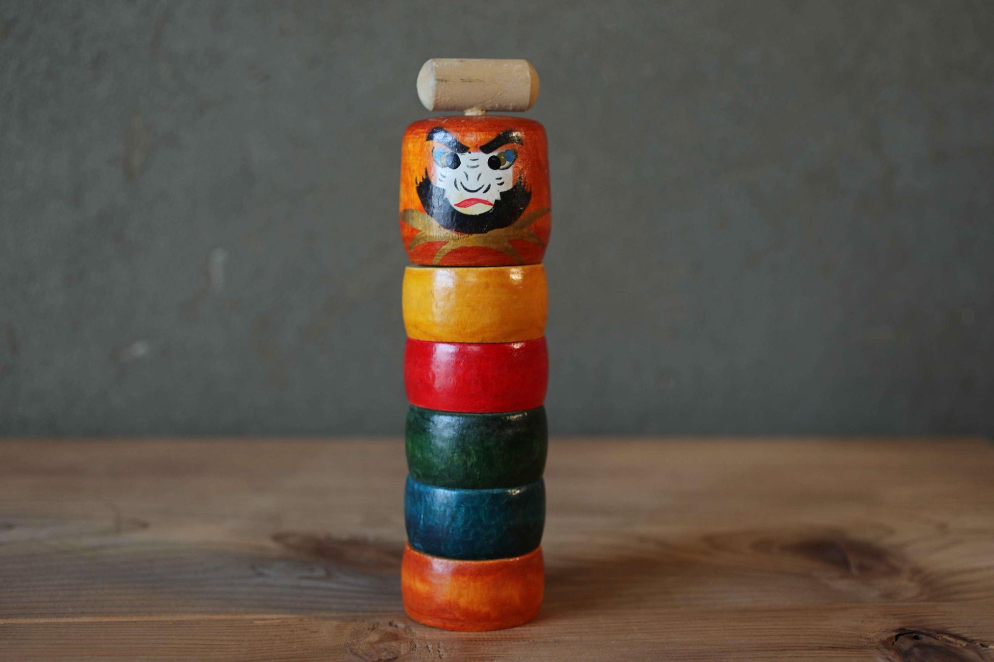 A colorful wooden toy with a face design sits on a wooden surface against a gray background.