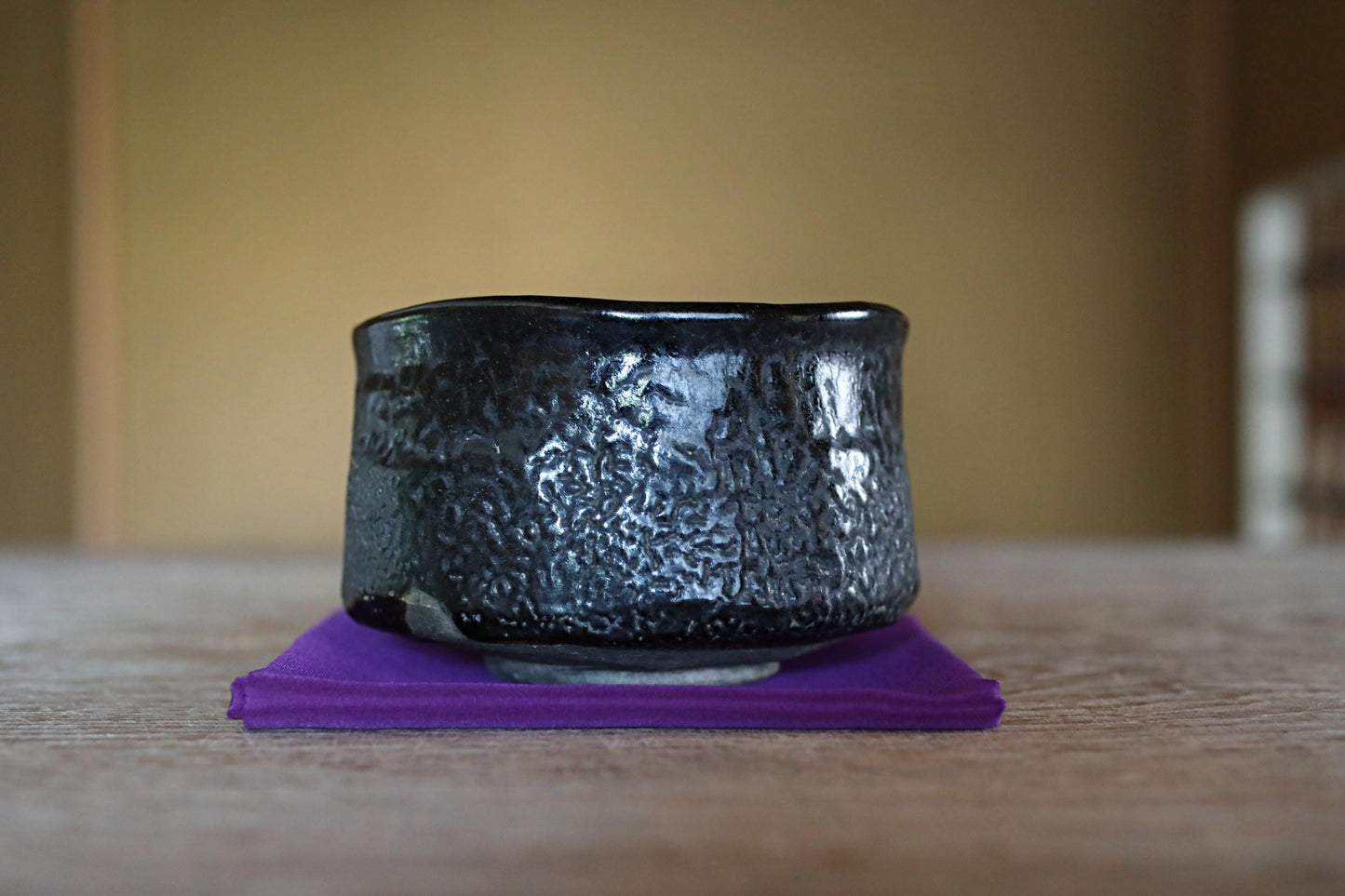 A black ceramic bowl with a textured surface sits on a purple cloth on a wooden surface.