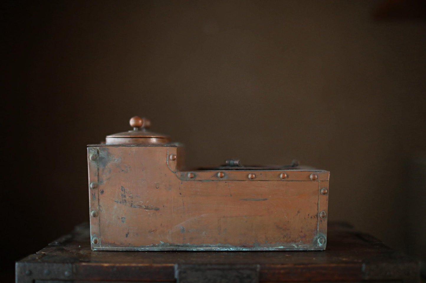 An old, weathered metal box with a lid, sitting on a wooden surface against a dark background.