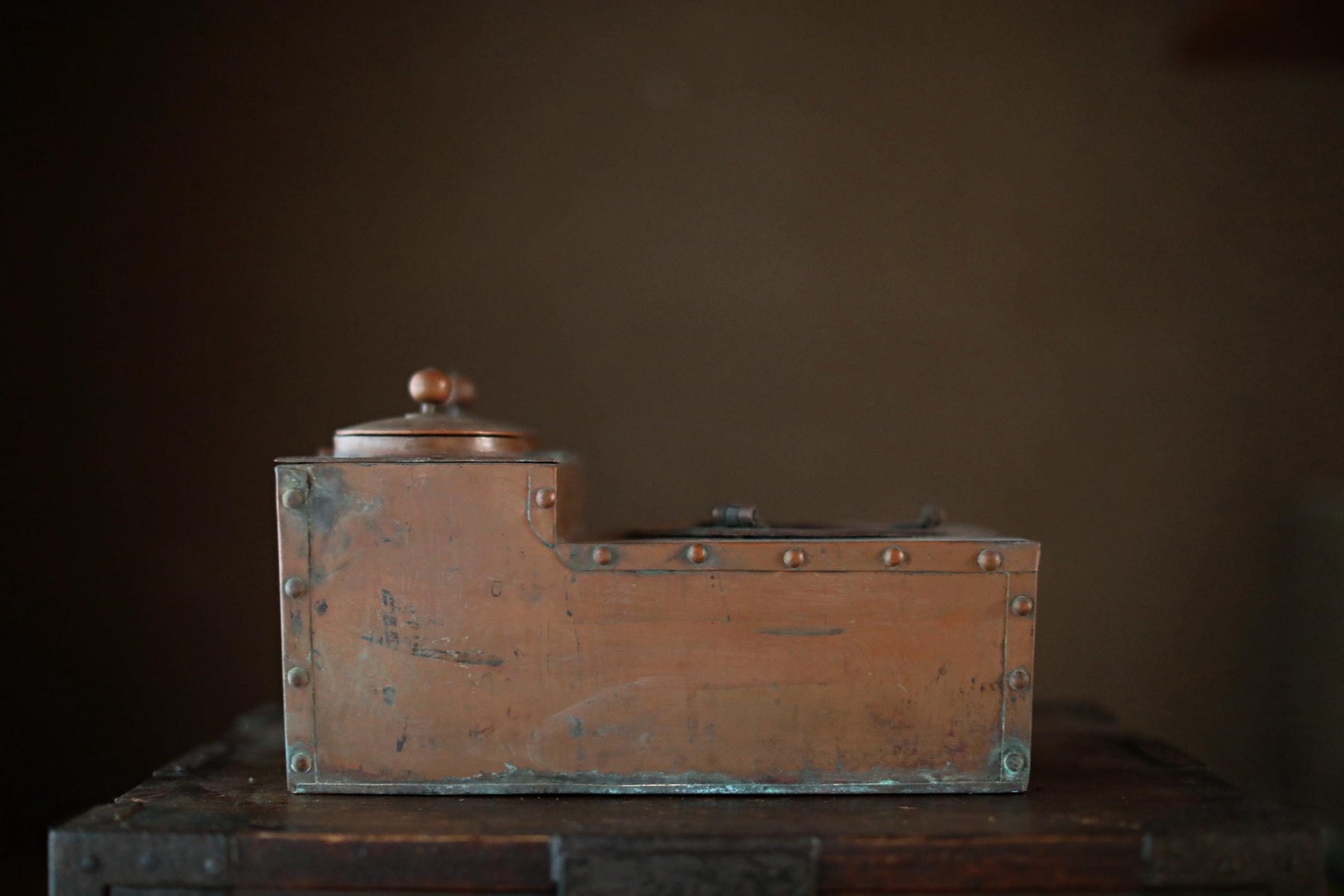 An old, weathered metal box with a lid, sitting on a wooden surface against a dark background.