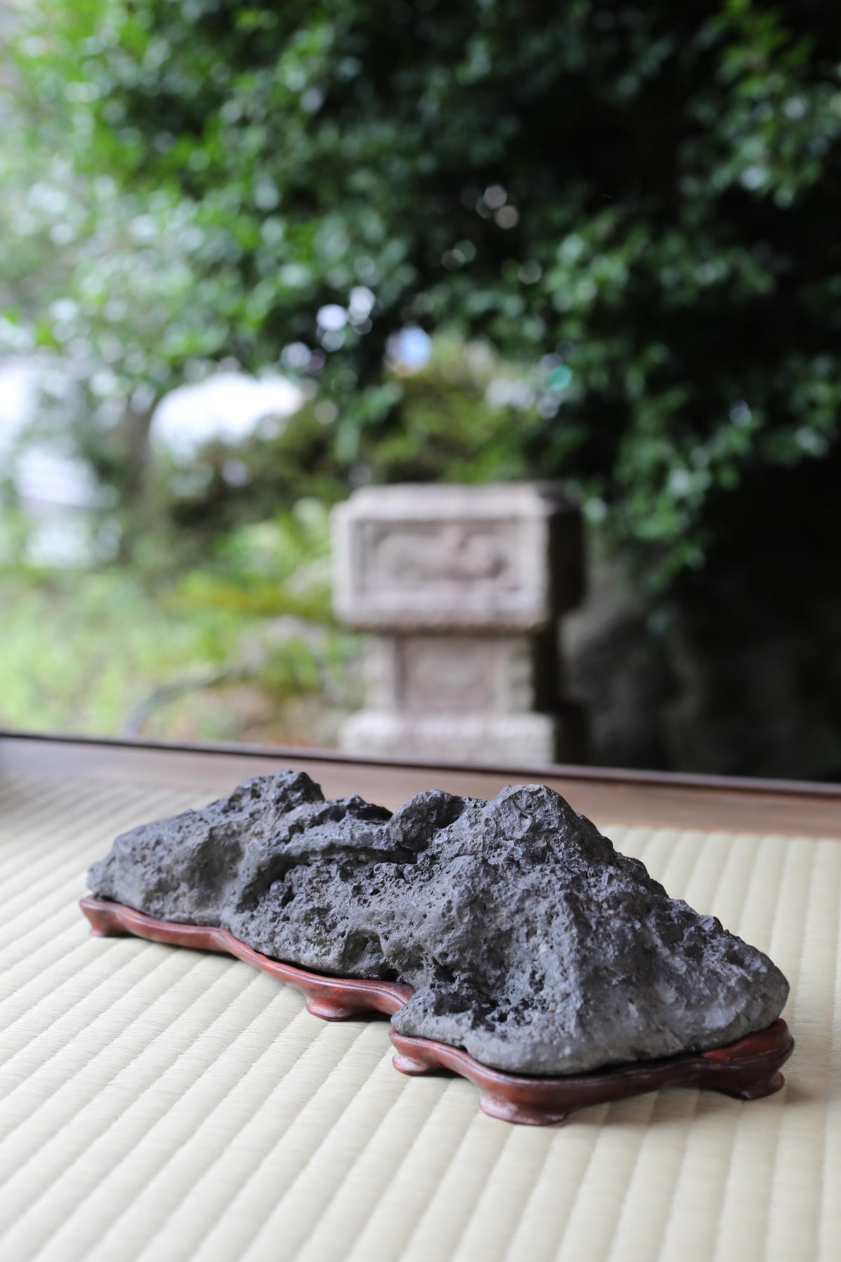 A large, dark gray rock sits on a wooden base, with a blurred natural background featuring trees and foliage.