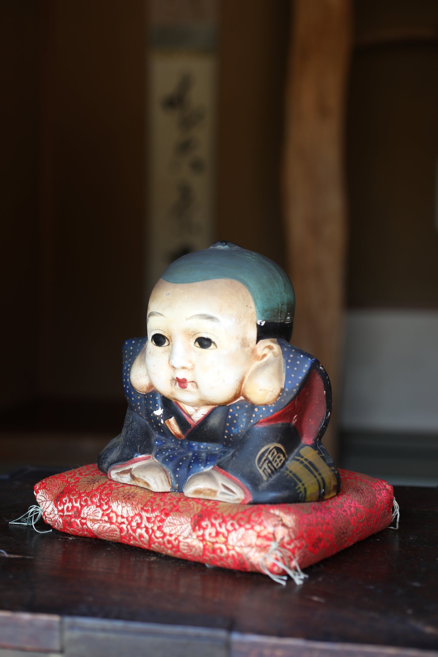 A ceramic figurine of a young boy wearing a blue and red outfit, sitting on a red cushion.