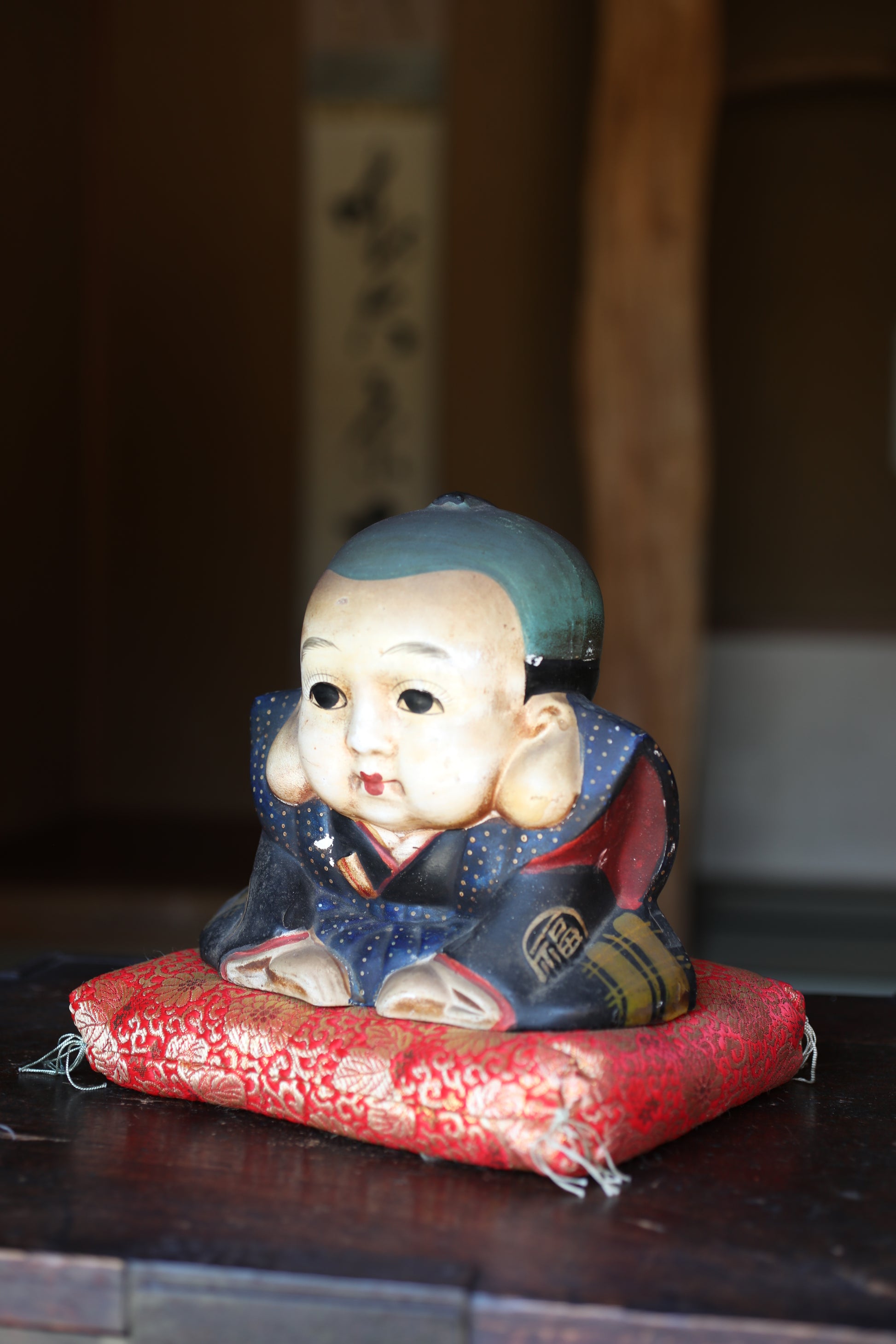 A ceramic figurine of a young boy wearing a blue and red outfit, sitting on a red cushion.