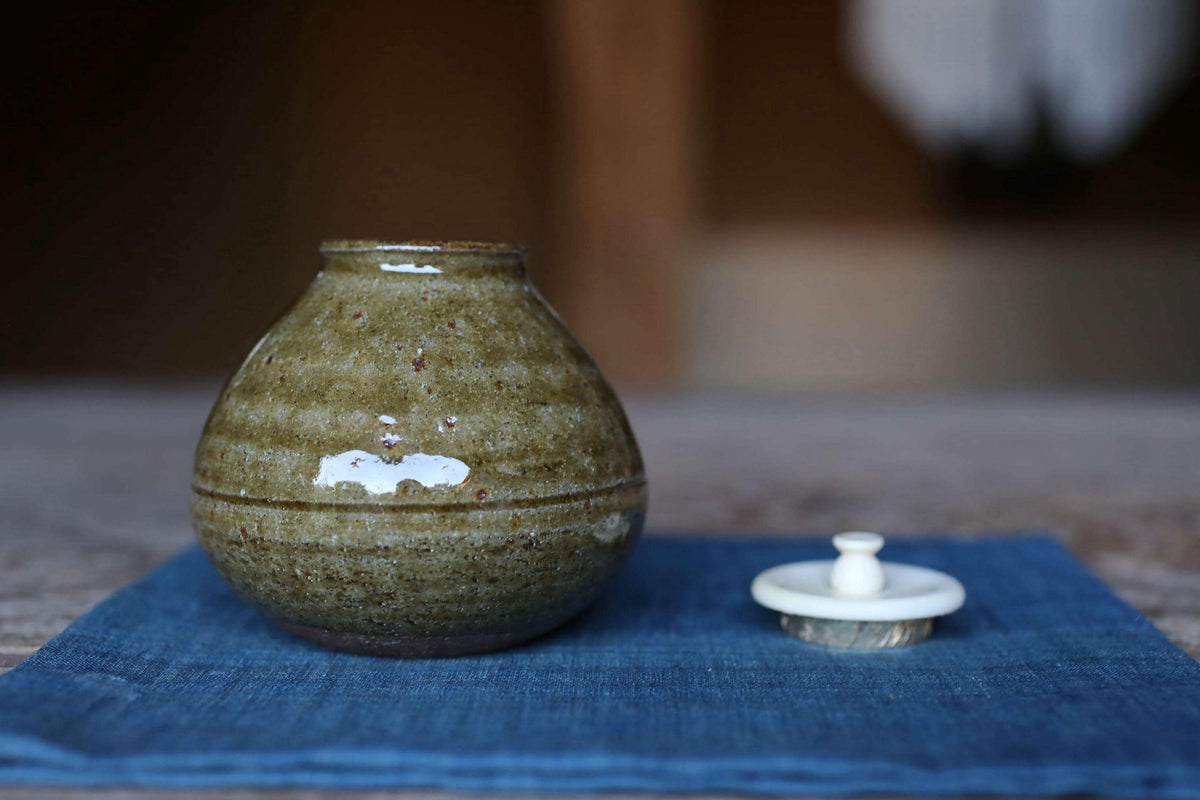 A ceramic jar with a white lid sits on a blue cloth, with a blurred background.