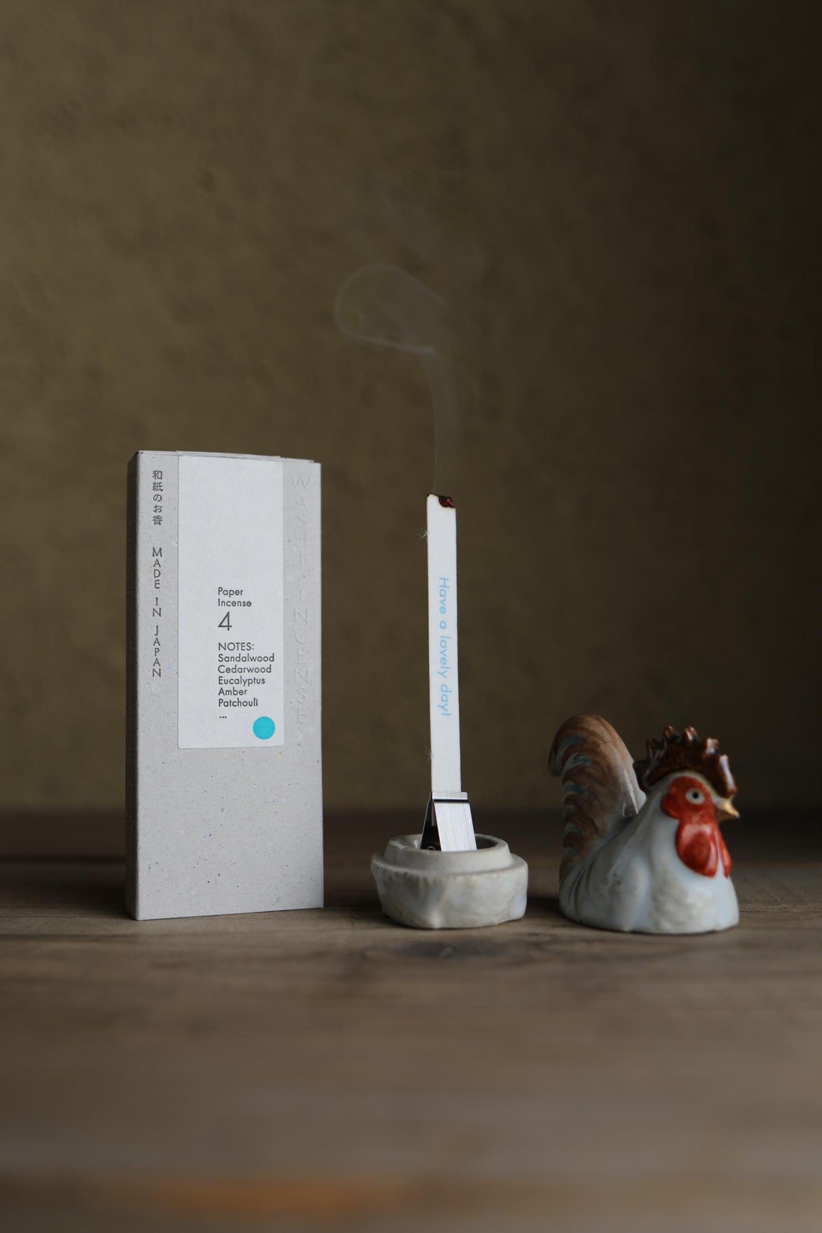 a white incense stick with smoke coming out of it, placed next to a ceramic rooster figurine and a box of incense sticks.