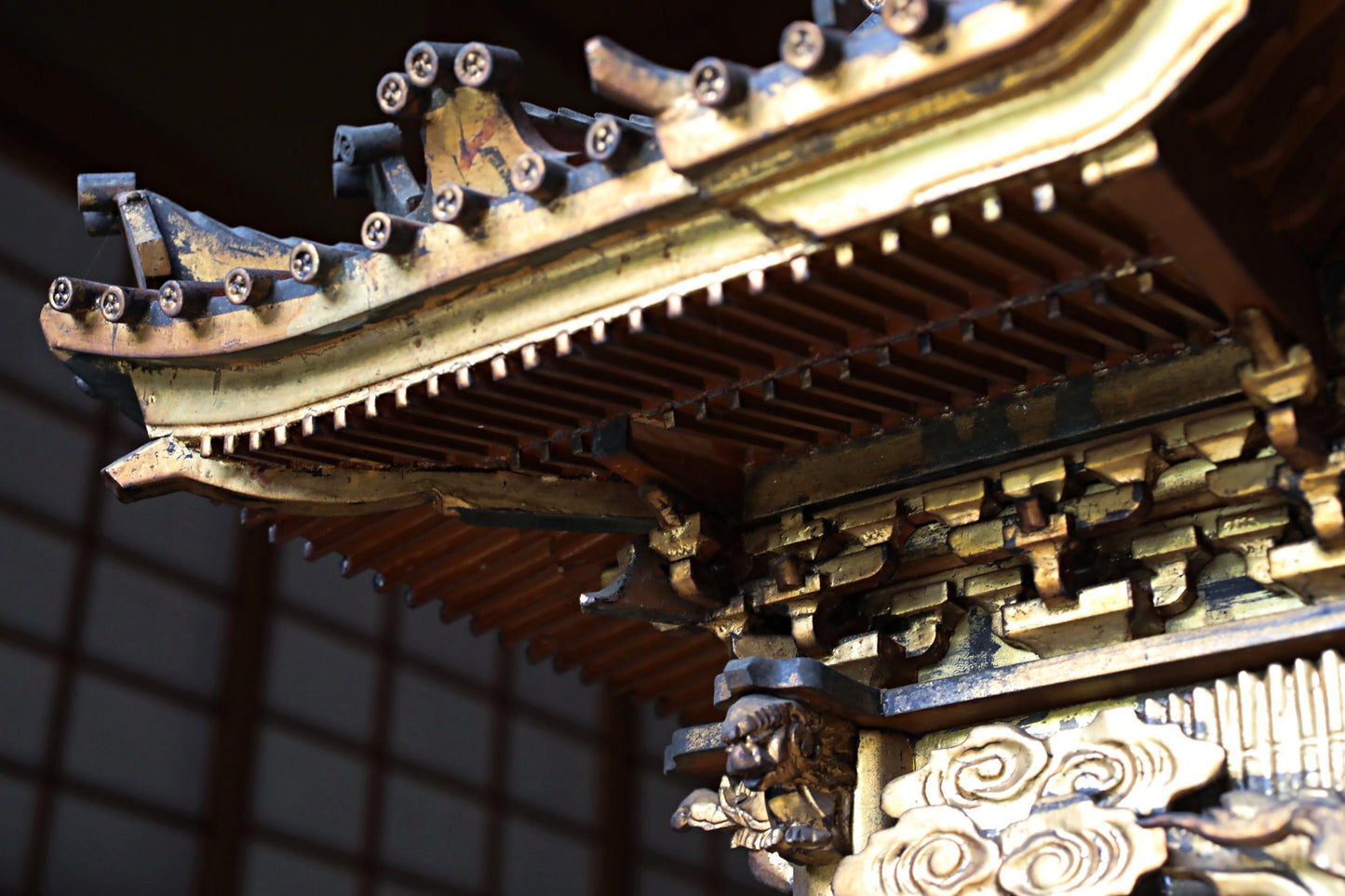 a close-up view of an ornate, intricately designed wooden structure, possibly a temple or a traditional architectural element. The structure features a detailed, decorative design with various elements such as a pointed roof, ornamental patterns, and a carved face. The image is focused on the intricate details and craftsmanship of the structure, highlighting its artistic and cultural significance.