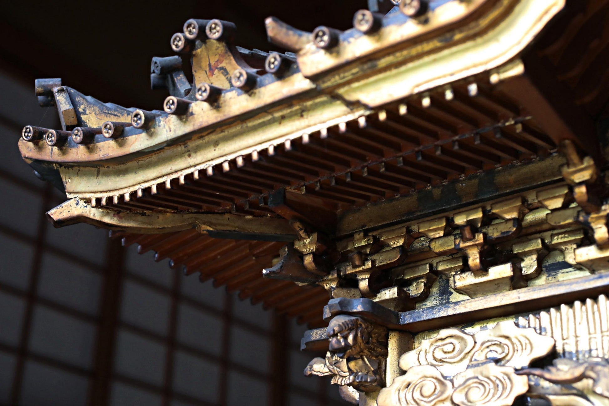 a close-up view of an ornate, intricately designed wooden structure, possibly a temple or a traditional architectural element. The structure features a detailed, decorative design with various elements such as a pointed roof, ornamental patterns, and a carved face. The image is focused on the intricate details and craftsmanship of the structure, highlighting its artistic and cultural significance.