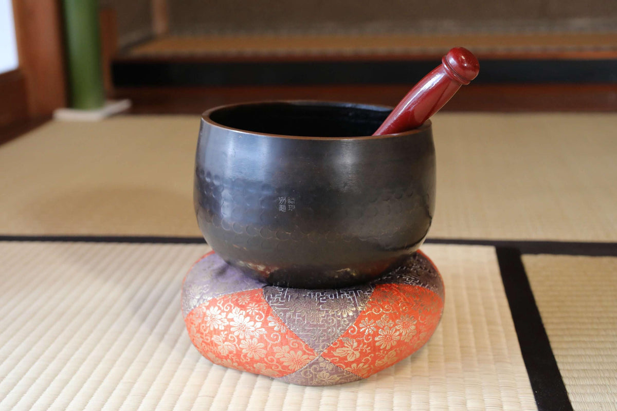A black bowl with a red handle is placed on a decorative orange and white cushion on a woven mat.