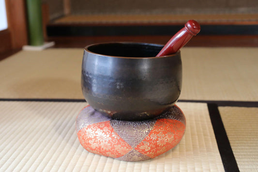 A black bowl with a red handle is placed on a decorative orange and white cushion on a woven mat.