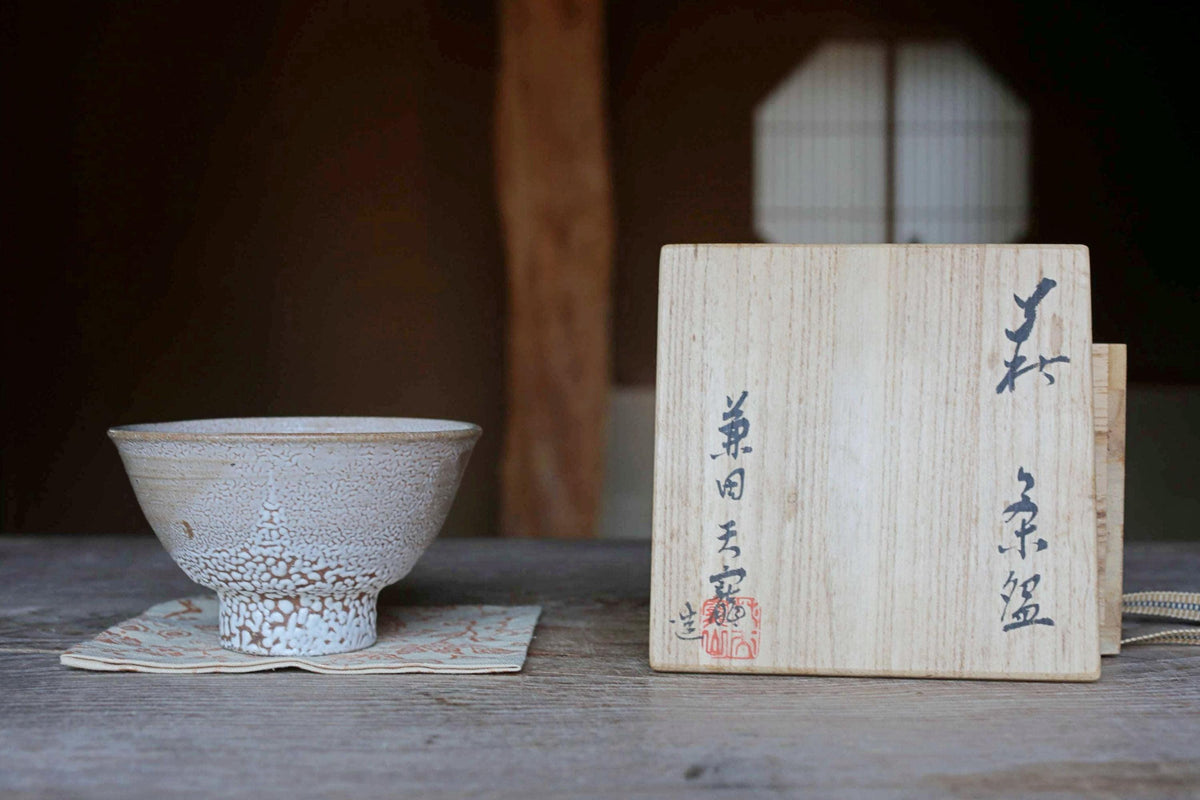 a wooden box with Japanese characters on it, placed next to a white ceramic bowl on a wooden surface.