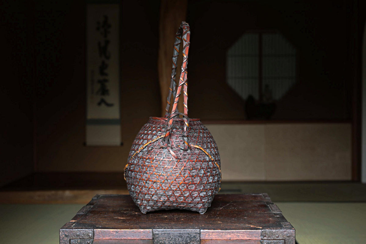 A traditional Japanese woven basket with intricate patterns sits on a wooden surface, with a window and a wall with Japanese calligraphy in the background.
