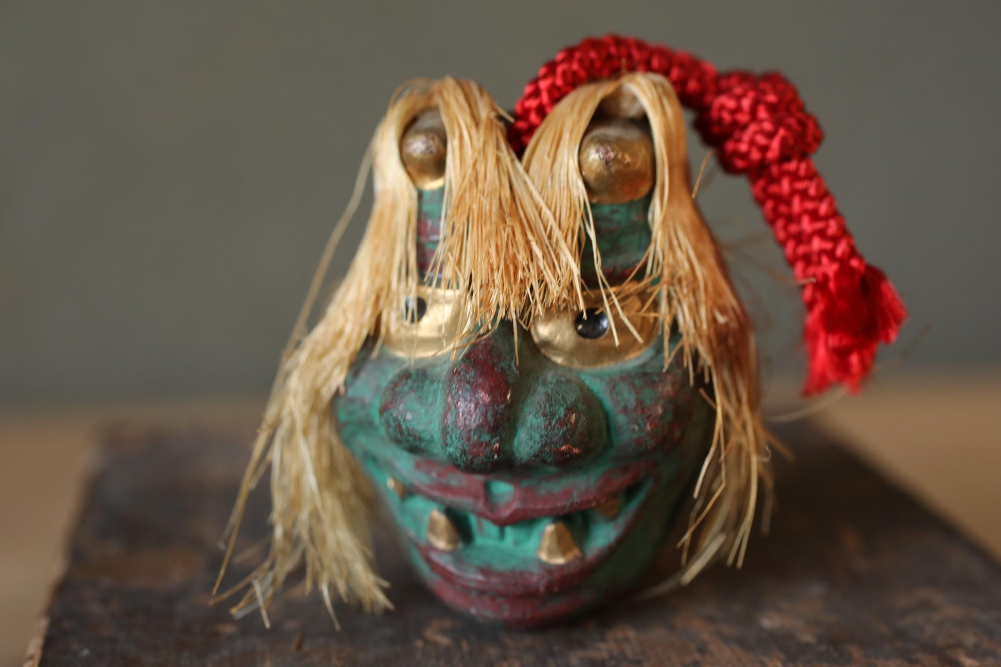 A colorful, whimsical mask with a red ribbon around its neck, sitting on a wooden surface.