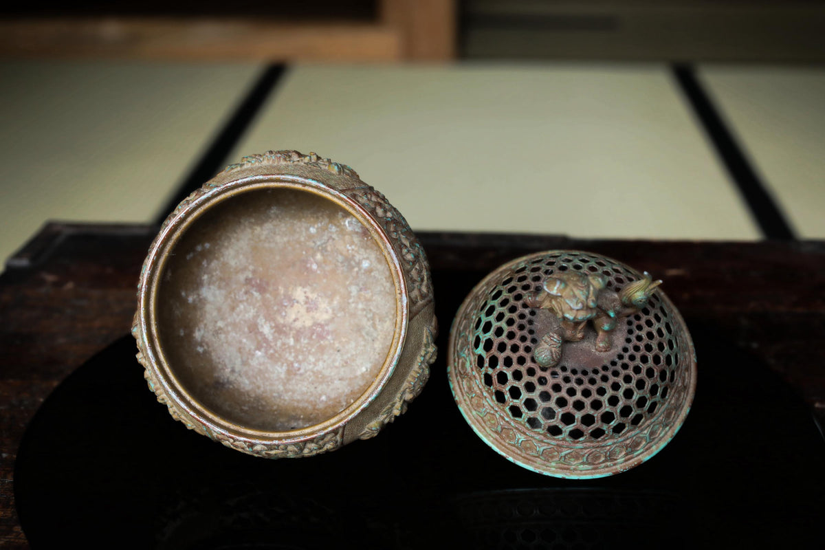An old, ornate metal box with a lid and a small, intricately designed lid, both sitting on a wooden surface.