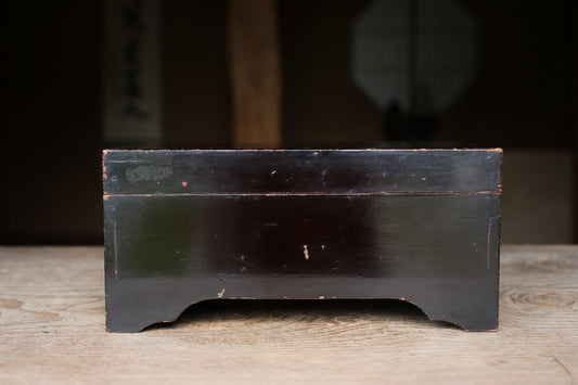 A black wooden box sits on a wooden surface, with a blurred background that includes a window and a wall.