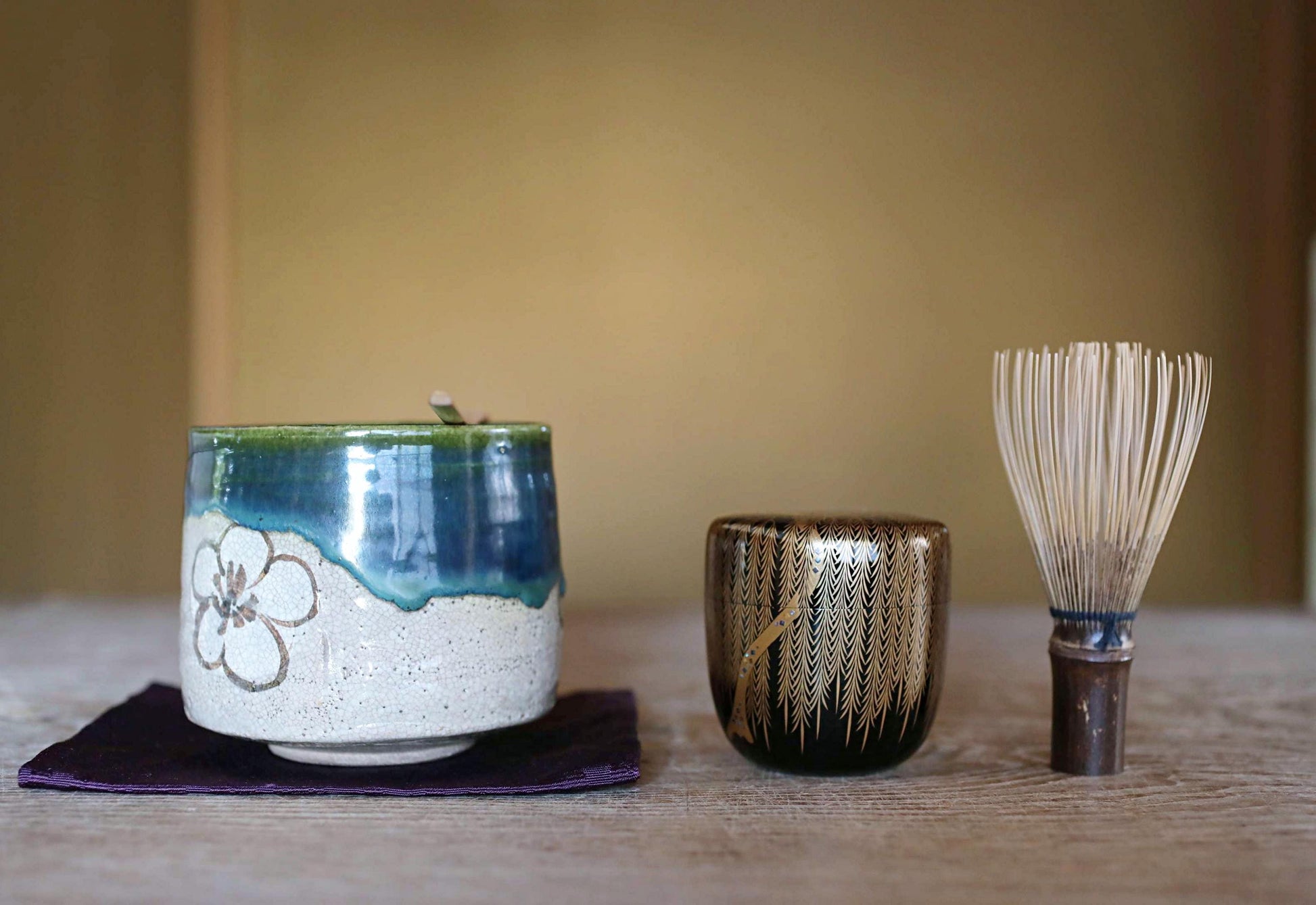 A ceramic teapot with a blue and white floral design, a small black and gold cup, and a white whisk are arranged on a wooden surface.