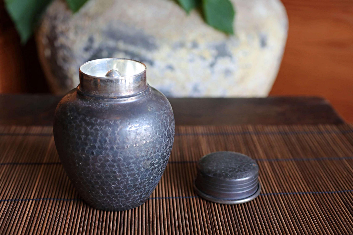 A black metal jar with a lid sits on a bamboo mat, with a blurred background that includes a vase and some greenery.