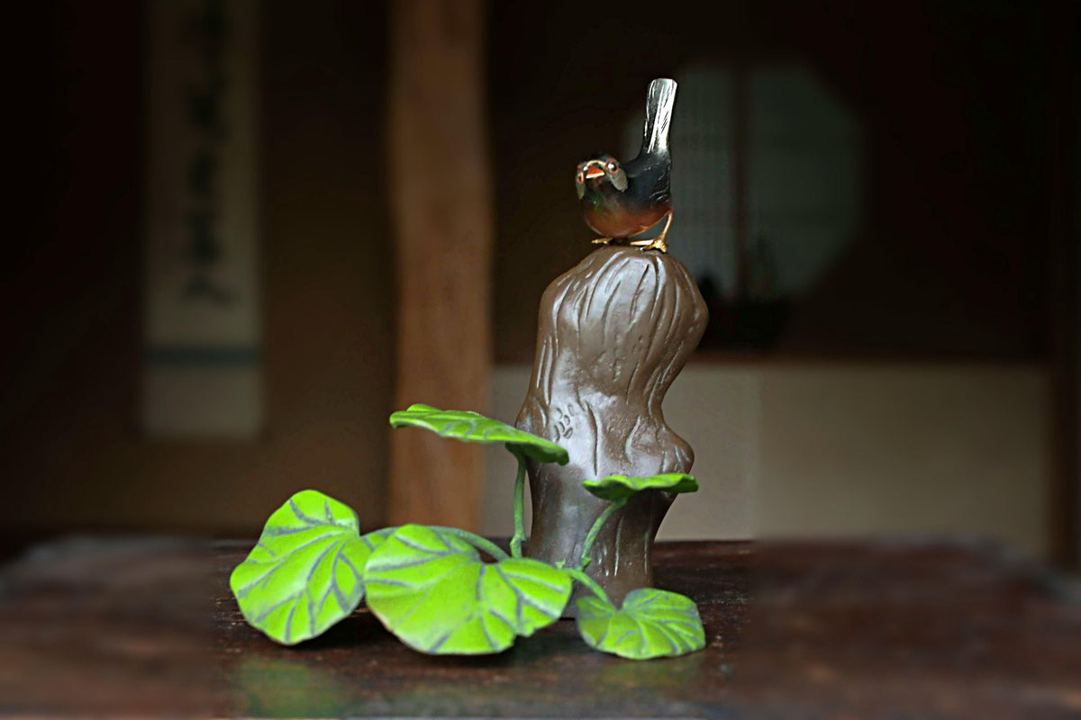 A ceramic bird figurine perched on a tree stump, surrounded by green leaves, sits on a wooden surface.