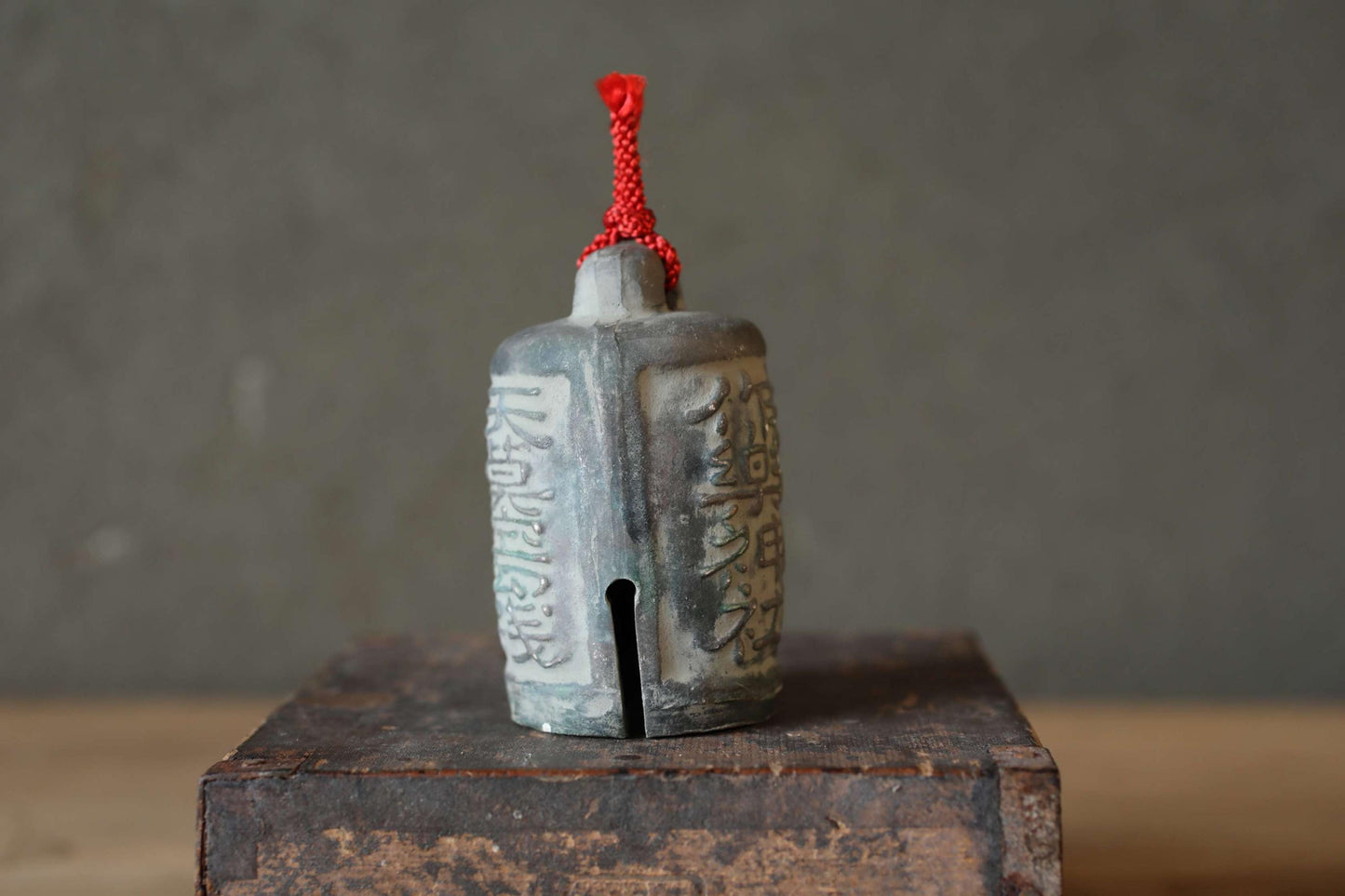 A small, gray ceramic bell with a red string hanging from the top, sitting on a wooden surface against a gray background.