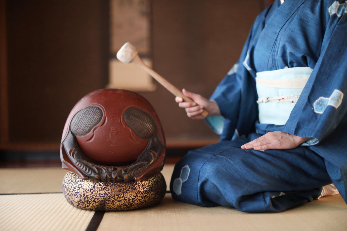A person in traditional Japanese attire is seated on the floor, holding a wooden object with a face on it, possibly a decorative item or a ceremonial object.