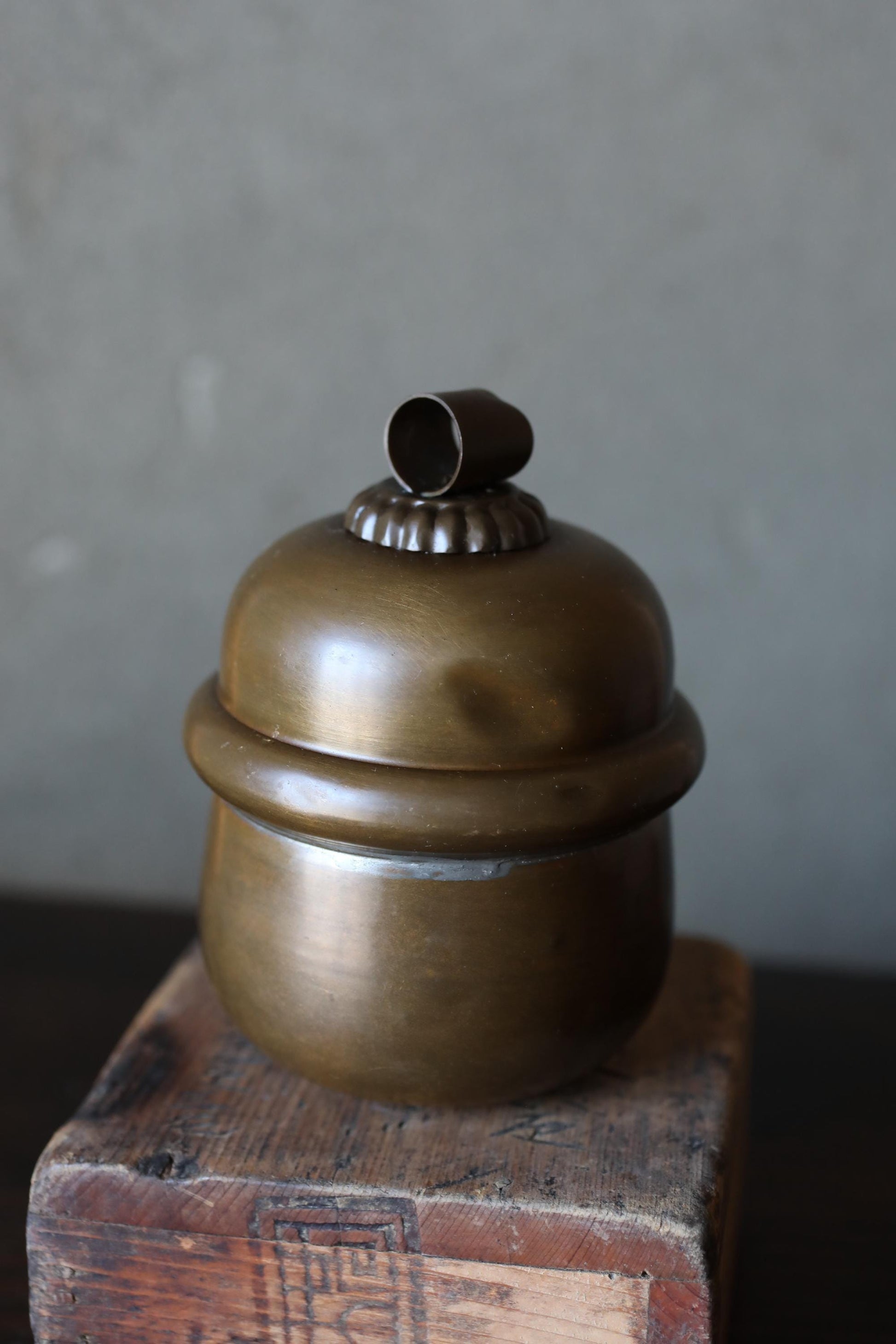 A brass-colored metal container with a lid sits on a wooden surface against a gray background.