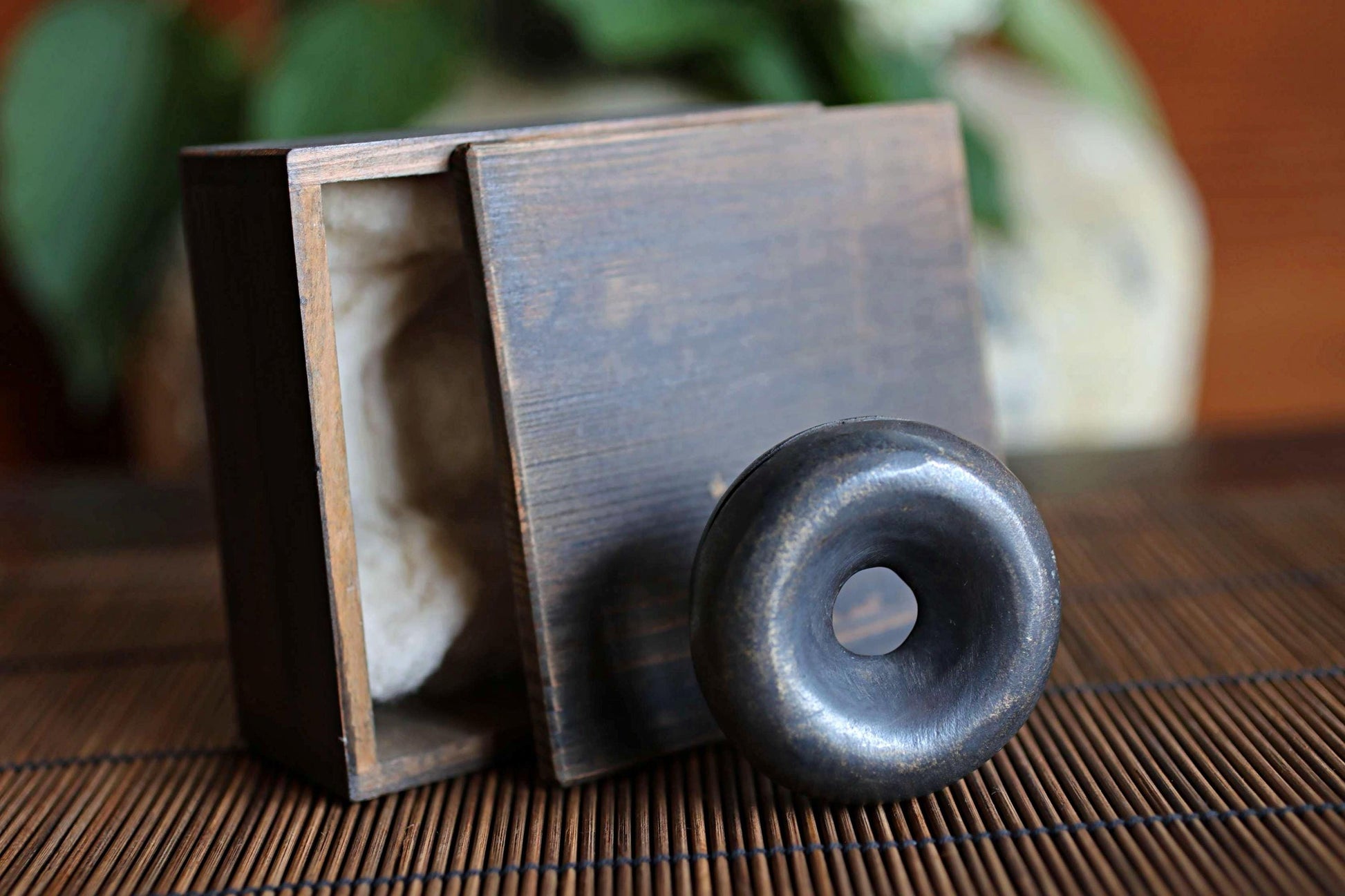 a wooden box with a lid, and a round metal object, possibly a sculpture or a piece of jewelry, placed on a bamboo mat.