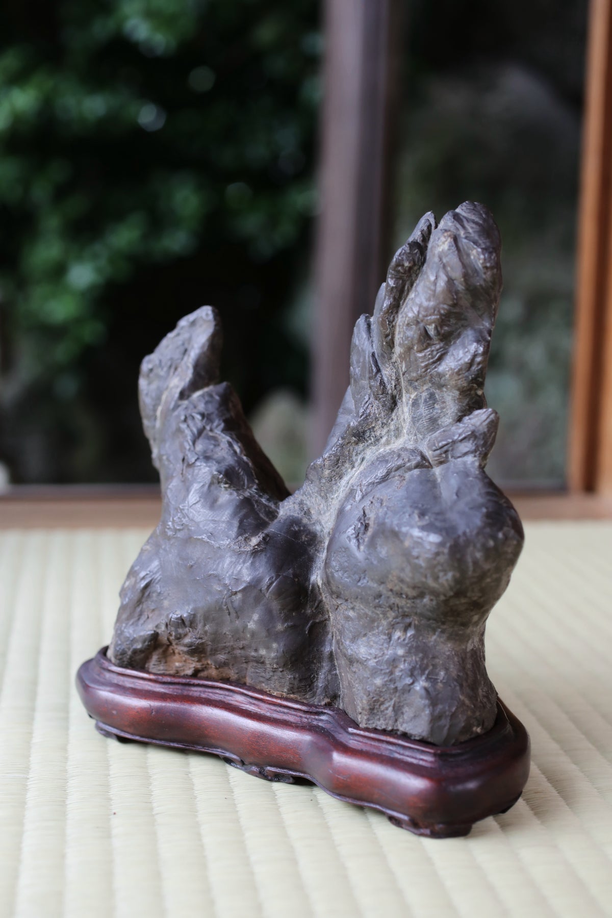 A dark gray stone sculpture with a wooden base is displayed on a woven mat.