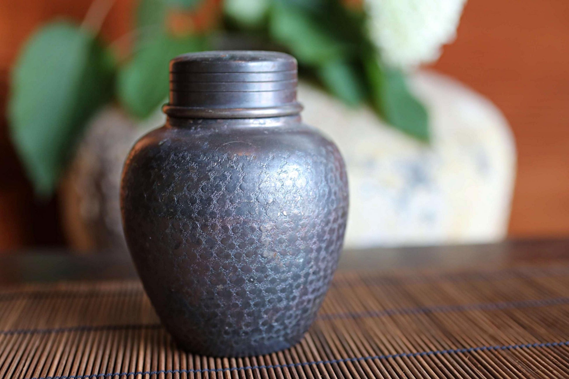 A dark-colored metal jar or container with a textured surface sits on a wooden surface, with a blurred background featuring a vase and some greenery.
