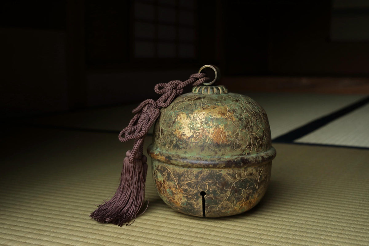 An ornate, antique-looking bell with a tassel hanging from it, placed on a woven mat.