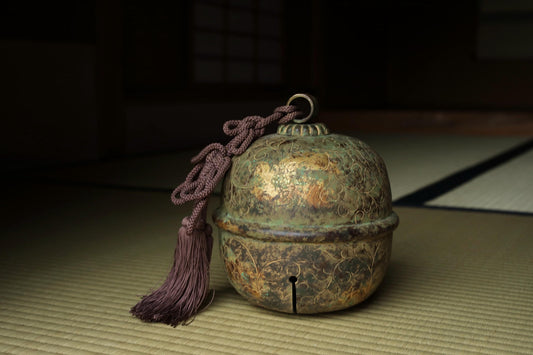 An ornate, antique-looking bell with a tassel hanging from it, placed on a woven mat.