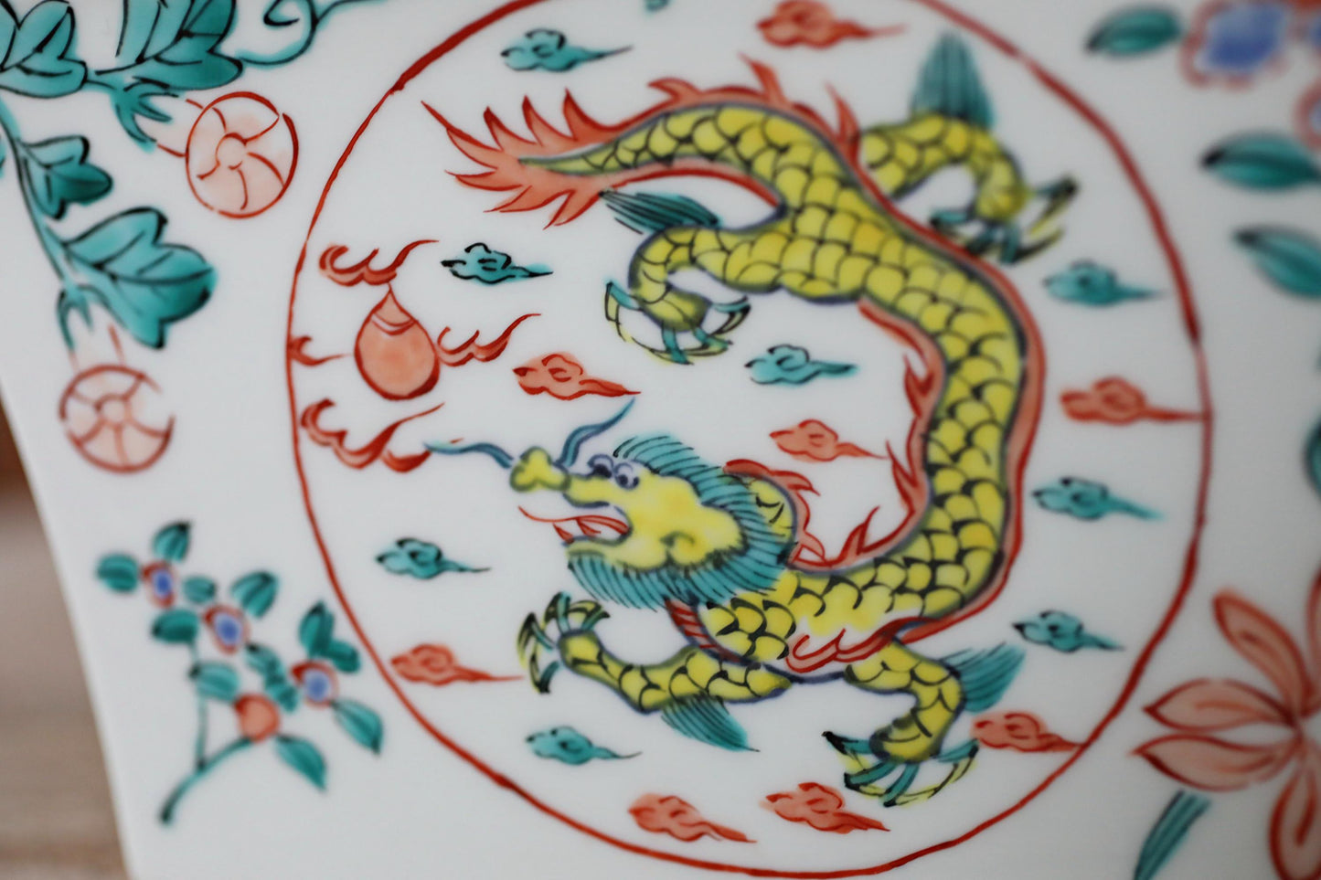 a close-up view of a decorative ceramic piece with a vibrant dragon design. The dragon is depicted in a dynamic pose, surrounded by various elements such as leaves, flowers, and clouds. The overall composition of the image is visually striking and captures the intricate details of the dragon design.