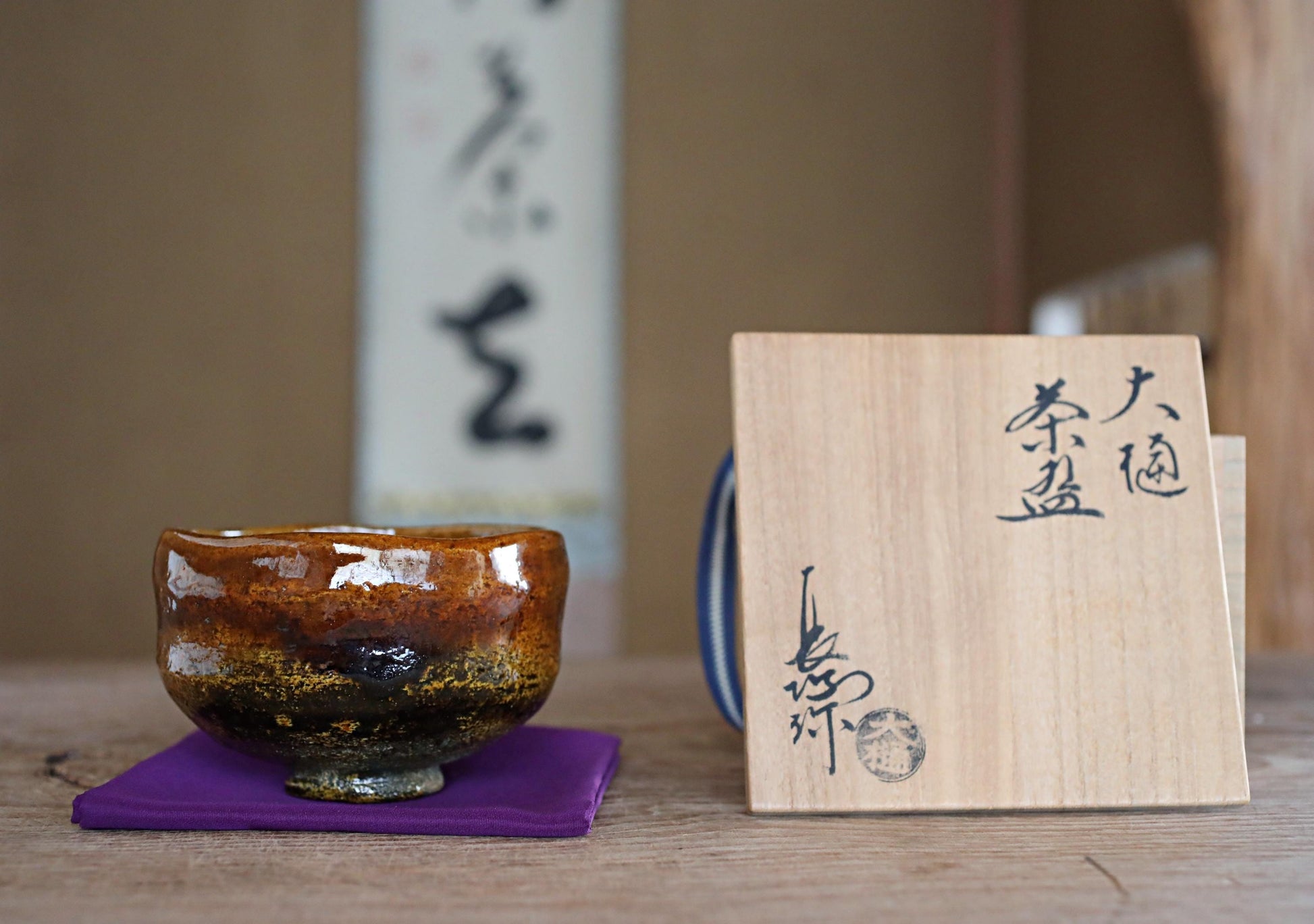 a brown ceramic bowl with a wooden chopstick placed on a purple cloth, with a wooden card featuring Japanese characters next to it.