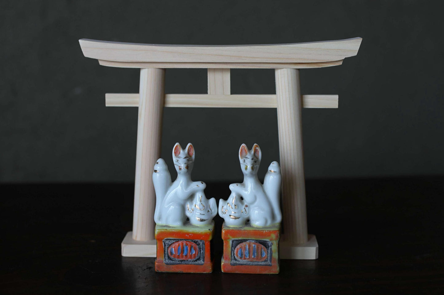 two ceramic figurines of cats, each sitting on a small orange and white box with the word "CHI" written on it. The figurines are positioned in front of a wooden torii gate, which is a traditional Japanese gate used at shrines and other sacred sites.
