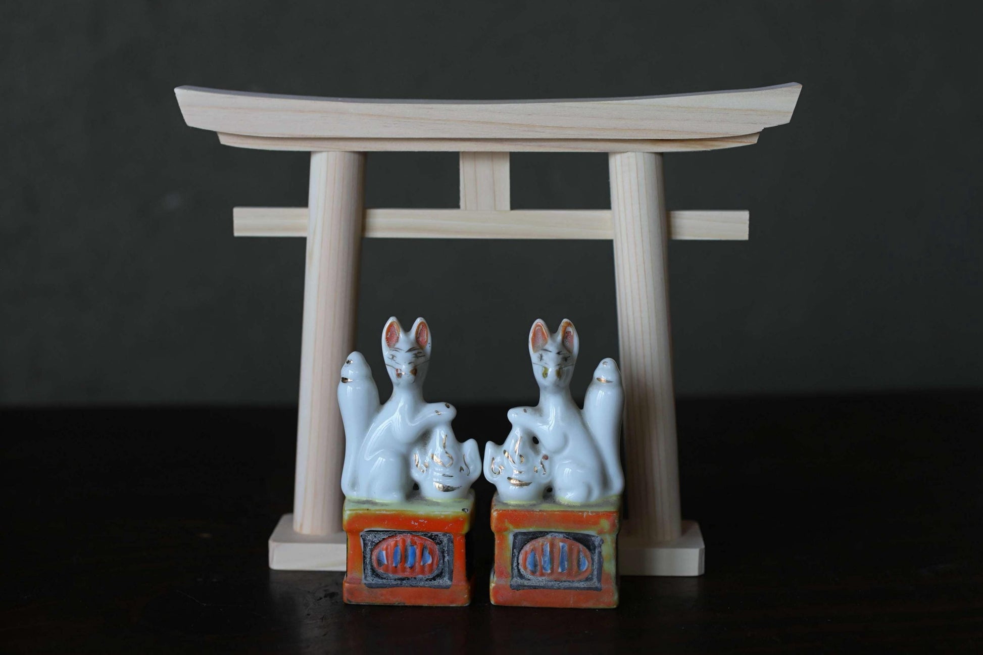 two ceramic figurines of cats, each sitting on a small orange and white box with the word "CHI" written on it. The figurines are positioned in front of a wooden torii gate, which is a traditional Japanese gate used at shrines and other sacred sites.