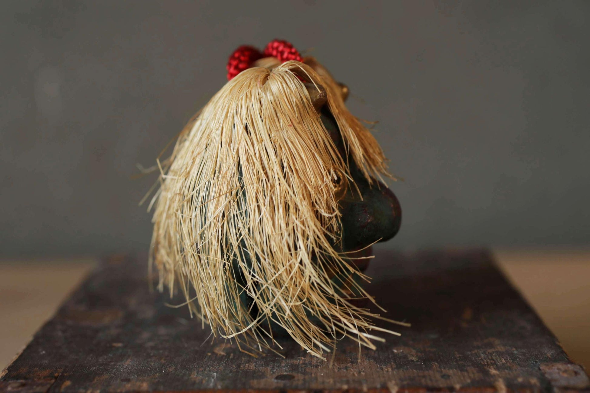 A small, black figurine with long, blonde hair and a red ribbon is displayed on a wooden surface against a gray background.