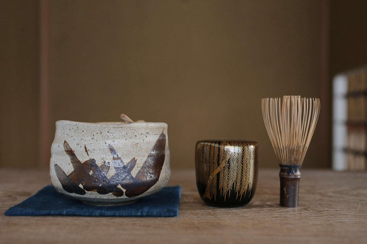 a wooden table with three objects on it: a white ceramic bowl with a black and brown design, a black and gold ceramic cup, and a wooden whisk.