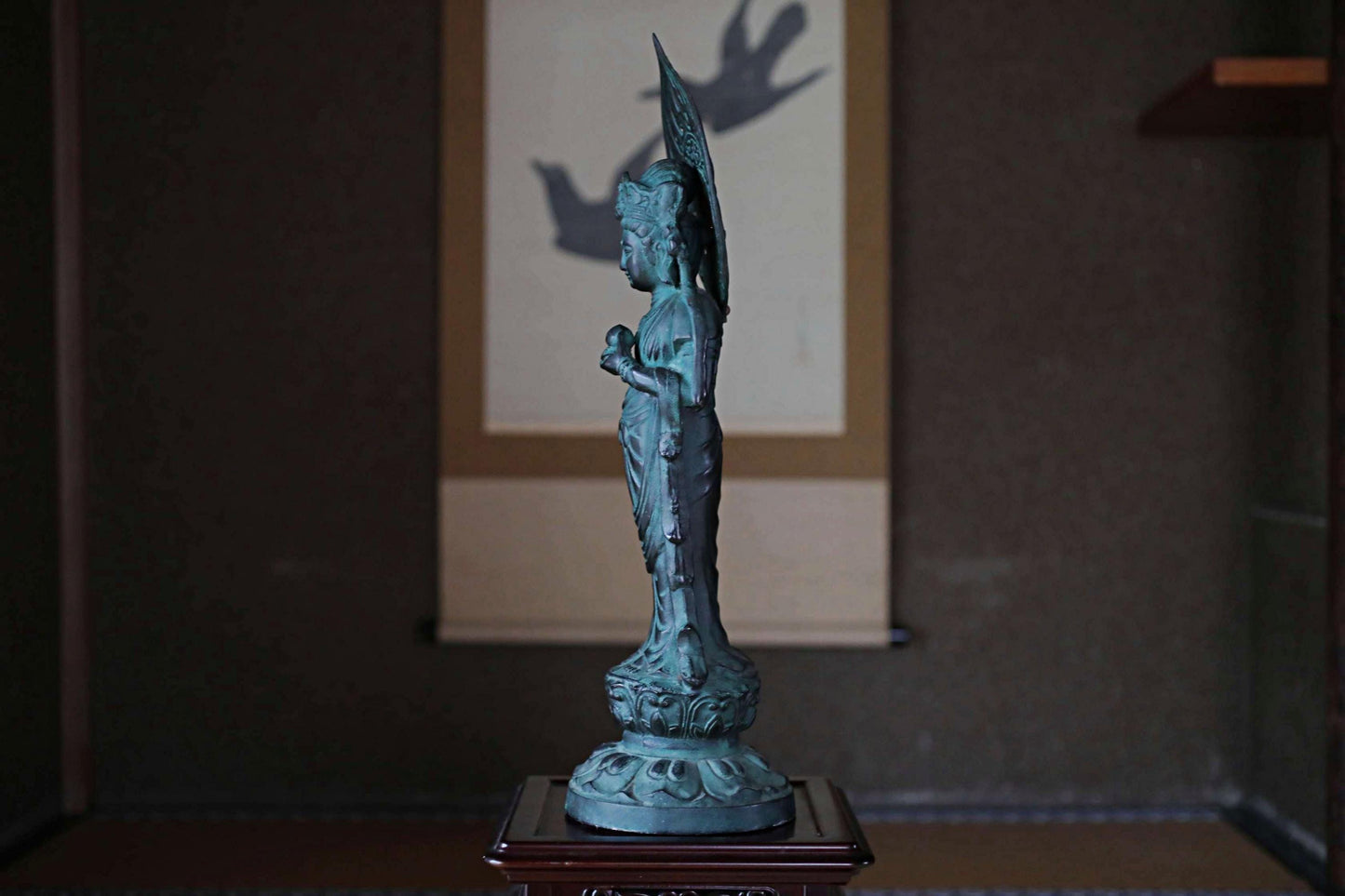A bronze statue of a deity stands on a wooden pedestal in a dimly lit room, with a painting of a deity visible in the background.