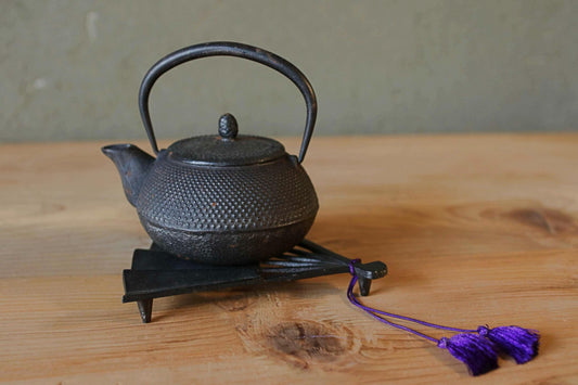 A black metal teapot with a curved handle and spout sits on a wooden surface, accompanied by a purple tassel.