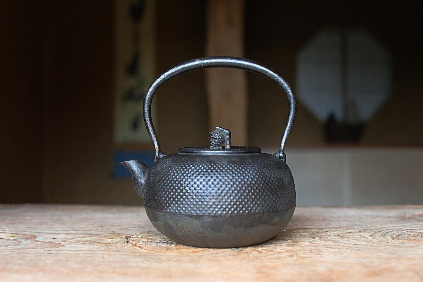 A black metal teapot with a handle and spout sits on a wooden surface, with a blurred background suggesting an indoor setting.