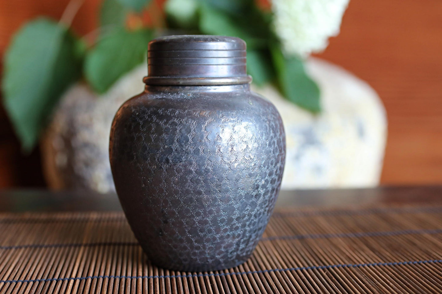 A dark-colored metal jar or container with a textured surface sits on a bamboo mat or placemat, with a blurred background featuring a vase and some greenery.