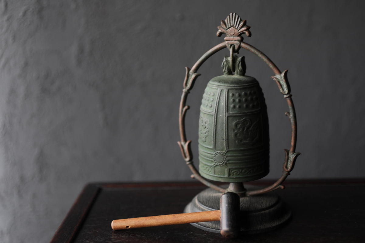 An antique green bell with a decorative metal frame sits on a dark surface, accompanied by a wooden mallet.