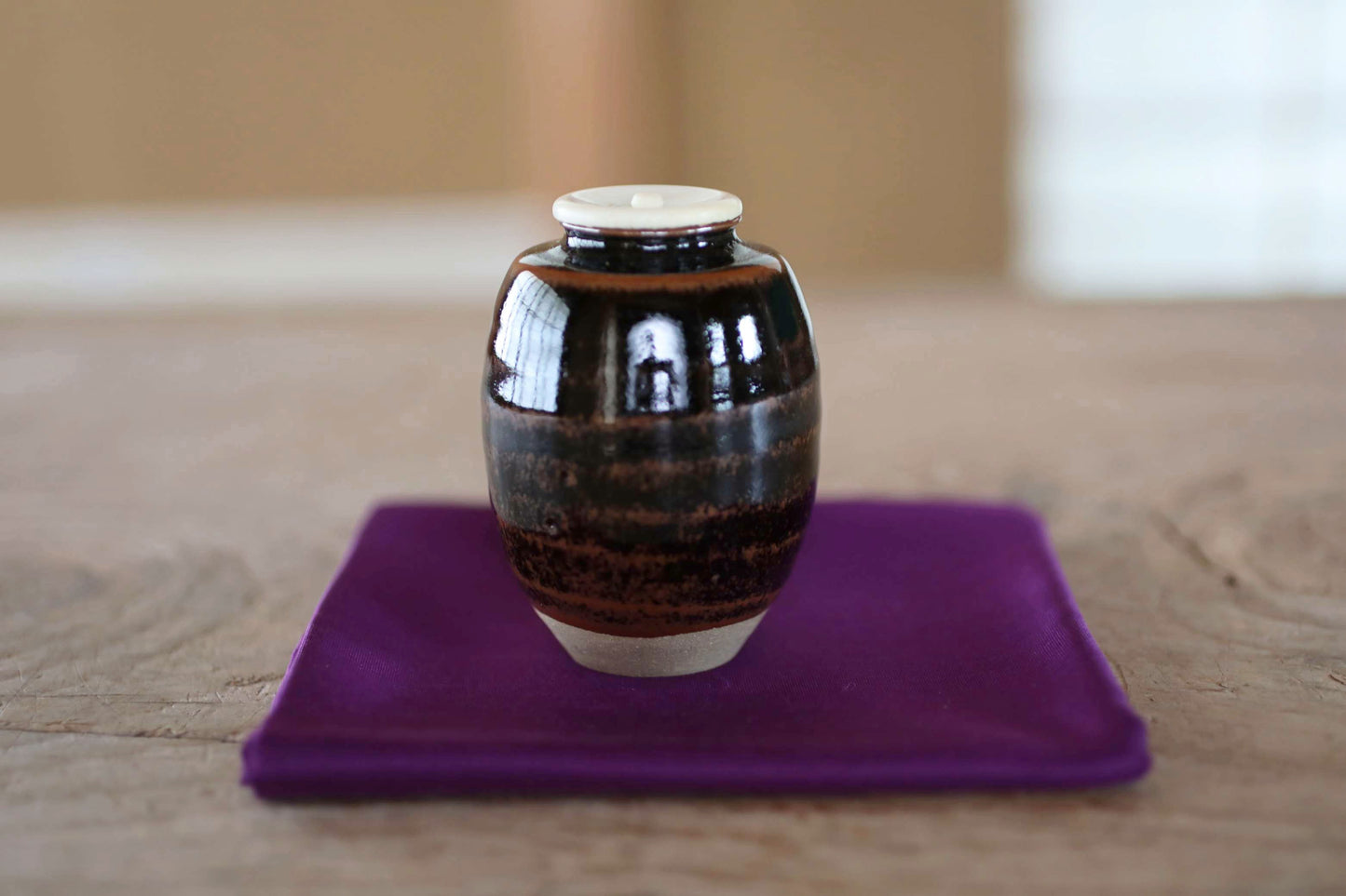 A small, round ceramic jar with a white lid sits on a purple cloth on a wooden surface.